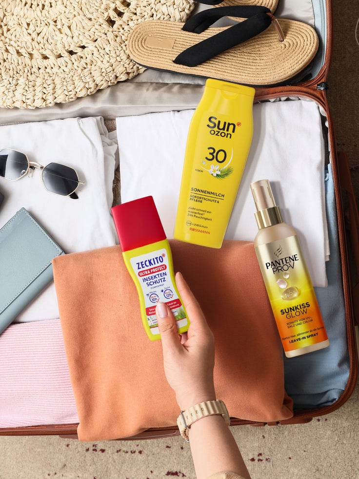 Geöffneter Koffer mit Kleidung, Sonnenhut, Sandalen, Sonnenbrille und Sommerpflegeprodukten, darunter Sonnenmilch, Insektenschutzspray und Haarpflege.