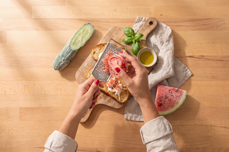Hände reiben gefrorene Tomate über Burrata auf geröstetem Sauerteigbrot, daneben liegen gefrorene Gurke, Wassermelone, Basilikum und Olivenöl auf einem Holzhintergrund.