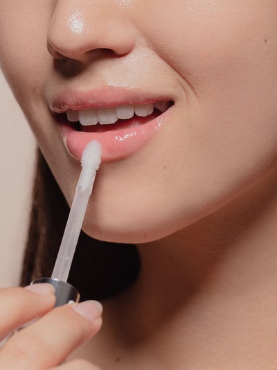 Lip Plumper auftragen: Nahaufnahme einer lächelnden Frau, die mit einem Applikator glänzendes, transparentes Lipgloss-Serum auf die Unterlippe aufträgt