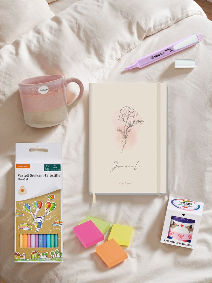 Flach gelegtes Arrangement für Journaling auf einem Bett: ein beigefarbenes Journal mit zarter Blumenillustration in der Mitte, daneben ein rosa Textmarker und eine Keramik-Tasse in Rosa-Beige. Unten liegen ein Set pastellfarbener Buntstifte, bunte Haftnotizzettel und eine Packung dekoratives Washi-Tape.