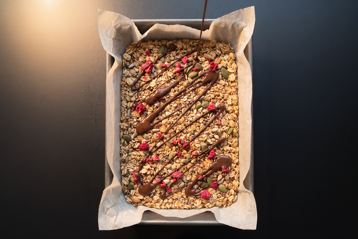 Müsliriegel-Masse mit Haferflocken, Kernen und Himbeeren wird in eine Form gepresst und mit geschmolzener Edelbitter Schokolade getoppt