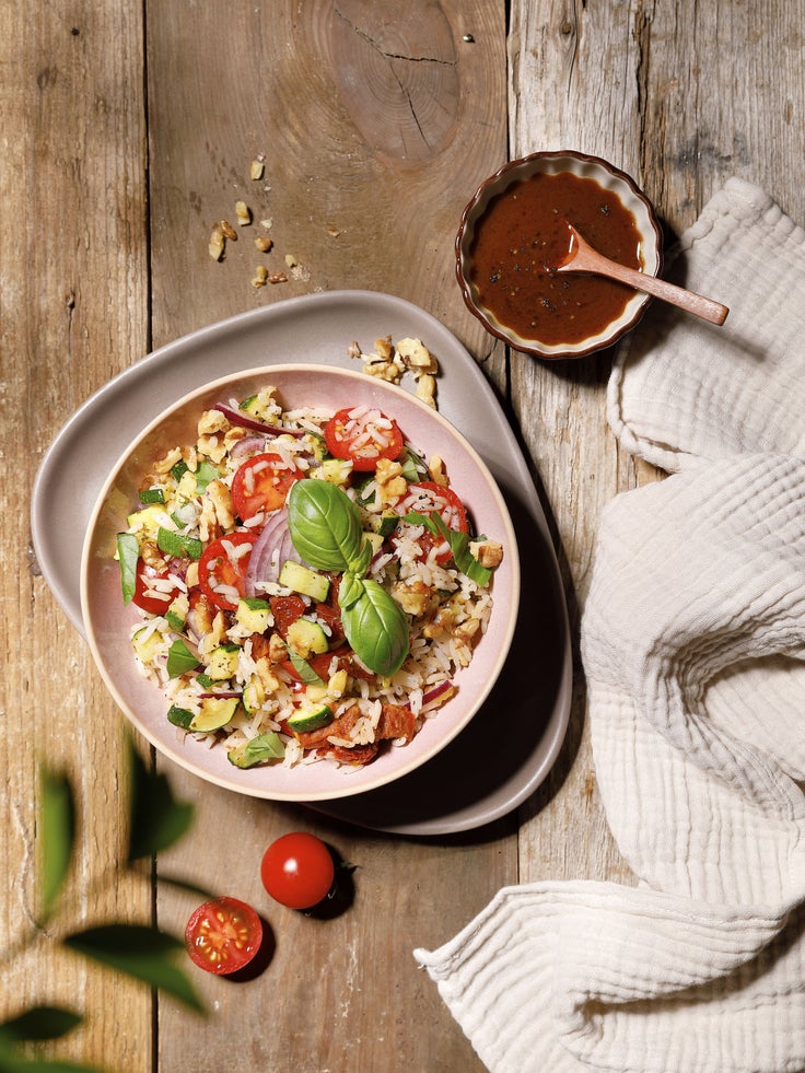 Schüssel mit sommerlichem Reissalat mit Tomaten, Zucchini, Basilikum und Nüssen auf Holztisch, daneben Dressing und helle Stoffserviette.