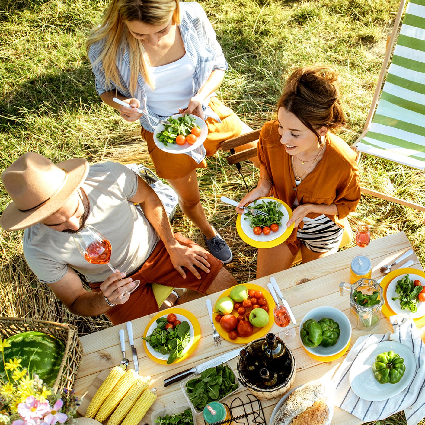 Sommerliches Picknick im Grünen mit drei Personen, buntem Gemüse, Salat und Getränken an einem Holztisch neben grün-weiß gestreiften Liegestühlen