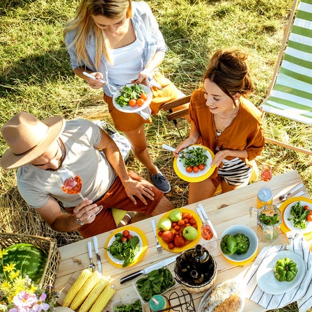 Sommerliches Picknick im Grünen mit drei Personen, buntem Gemüse, Salat und Getränken an einem Holztisch neben grün-weiß gestreiften Liegestühlen