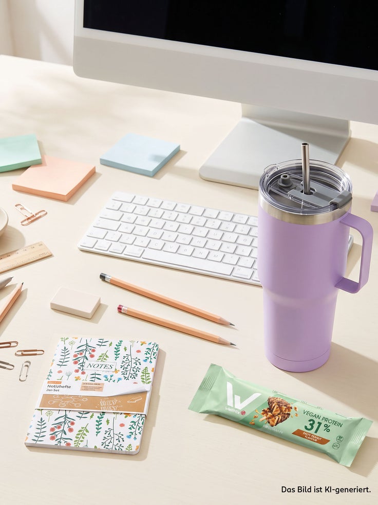Heller Schreibtisch mit Tastatur, Notizbüchern, Stiften, einem lilafarbenen Trinkbecher und veganem Proteinriegel.